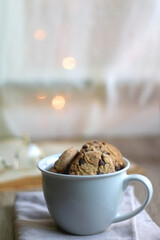Cup of chocolate chip cookies, open book and reading glasses on the table. Lit candle and bokeh lights in the background. Hygge at home. Selective focus.