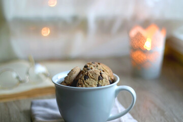 Cup of chocolate chip cookies, open book and reading glasses on the table. Lit candle and bokeh lights in the background. Hygge at home. Selective focus.
