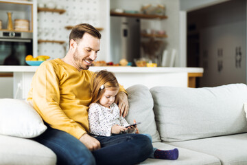 Single father and his small daughter using cell phone on sofa at home