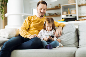 Single father and his small daughter using cell phone on sofa at home