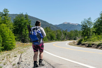 una mujer caminando viajando con la mochila en una ruta con arboles y montaña en verano