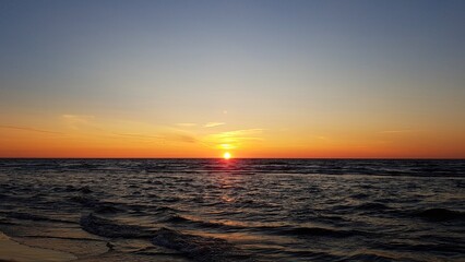 The sun in cloudless sky going beyond the horizon of the darkening sea