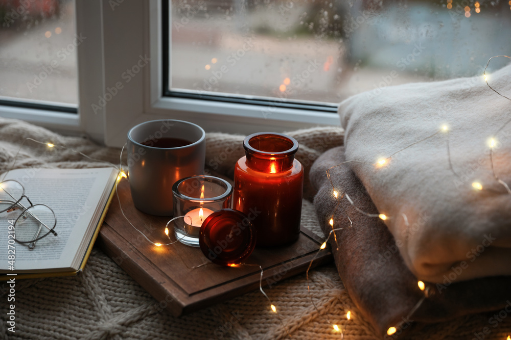 Sticker Cup of hot drink, open book and Christmas lights on knitted blanket near window. Cozy season