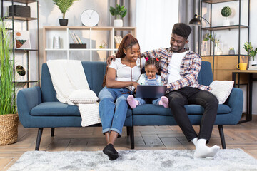 Obraz premium Happy african american parents with cute daughter using wireless laptop while sitting on couch. Family watching cartoons during leisure time at cozy apartment.