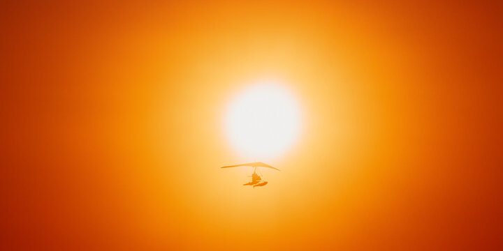 Motorized Hang Glider Flying In Clear Sunny Sunset Sky With Sun On Background.
