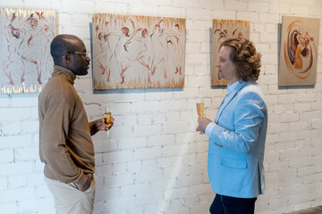 Two men standing near the wall with paintings drinking champagne and discussing modern art