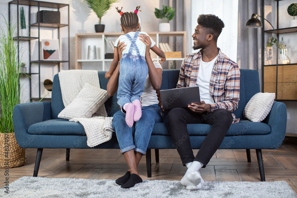 Wall mural African american mother playing with her little daughter on couch while smiling father sitting near with modern laptop on knees. Technology, family and leisure time concept.