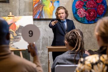 Mature man in suit pointing at man with tablet, people buying paintings of famous artists during auction in gallery - obrazy, fototapety, plakaty