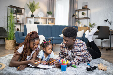 Beautiful and happy african american family drawing with colorful pencils during leisure time at cozy home. Concept of children, parents and education.