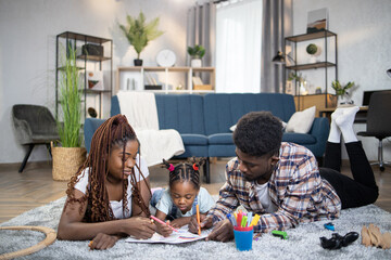African american parents resting on floor with their little daughter and drawing with pencils in album. Leisure activity of young family at home.