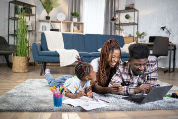 African married couple smiling cheerful while looking on computer screen. Little daughter drawing with colorful marker in album. Happy family lying on floor.