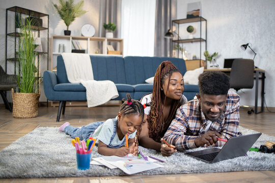 Happy african parents using modern laptop while their little daughter drawing in album. Young family of three lying on floor and enjoying time at home.