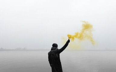 Male teenager in black clothes holding colorful yellow smoke sticks up in the air over moody gray...