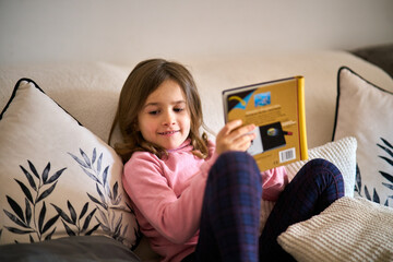 A little girl sitting on a couch and reading a book.