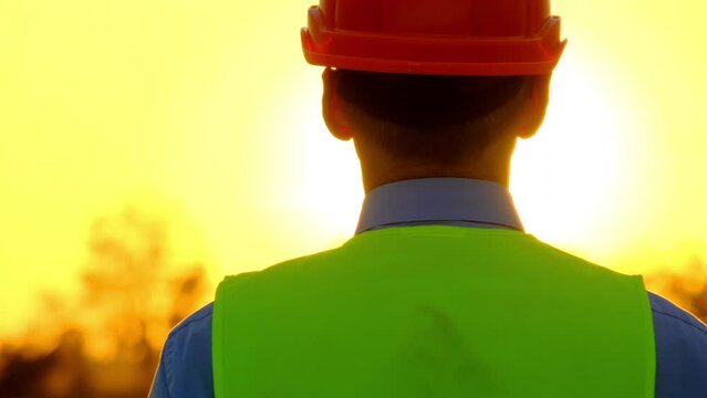 Engineer at vivid sunset. Specialist of maintenance in orange hardhat walks towards huge bright sun on rural windmill station backside view