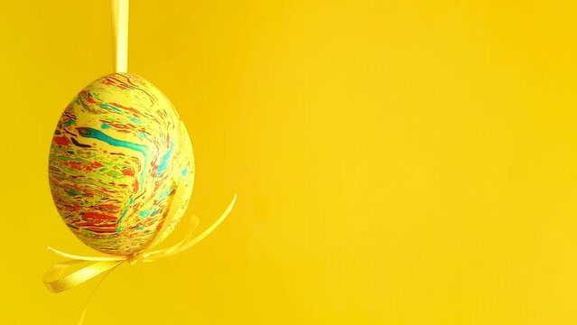 Easter egg of yellow green and orange colors hanging in the air on the left side of the frame on blurred orange background. Spring family holidays and decoration close up concept with copy space