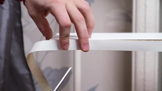 A man glues white melamine edging to the raw edge of wood chipboard furniture.