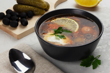 Traditional russian meat soup Solyanka in a black bowl and ingredients on concrete grey background