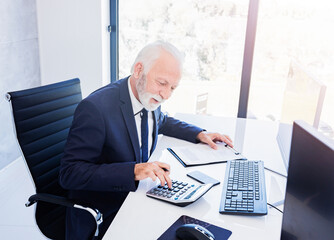 Gray haired businessman with beard in suit sitting at work desk and using calculator.