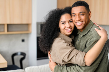 Close-up of happy multiracial couple posing while standing at the kitchen with blurred background. Happy owners of new flat smiling and embracing in front of the camera