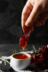 Woman dipping spicy beef jerky into sauce in bowl on dark background