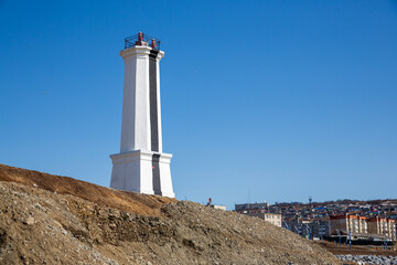 Lighthouse (leading beacon) on the sea coast in the port town. White lighthouse against the blue sky. Marine navigation and infrastructure. Magadan, Magadan Region, Far East of Russia.