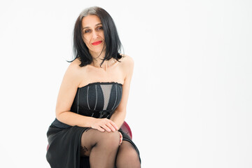 Fototapeta premium Middle aged woman starting getting grey-haired is posing in studio while sitting on the chair in black dress and stockings on white background, middle age skincare cosmetics, cosmetology concept