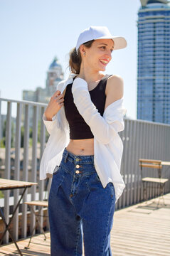 Female portrait of young active girl in black top, white shirt, basketball cap, and jeans on modern buildings background outdoors