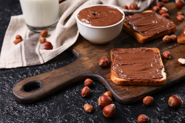 Wooden board of bread with chocolate paste and hazelnuts on black background
