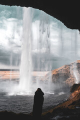 Vertical View of Seljalandsfoss Waterfall in Iceland from behind waterfall, with a silhouette of a woman in a fur hood jacket