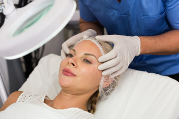 Specialist is analysing skin of female patient with using magnification before the procedure in clinic.