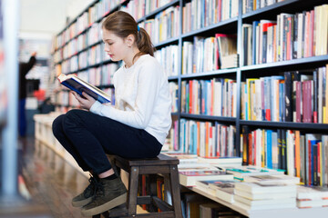 Cute little glad positive girl absorbed in reading book while sitting on small wooden ladder in...