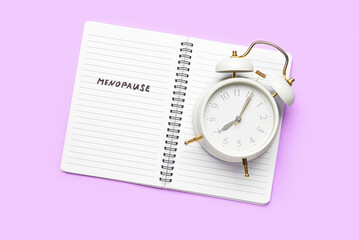 Notebook with word MENOPAUSE and alarm clock on lilac background