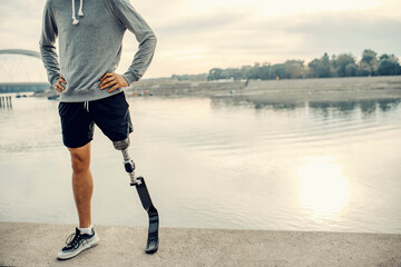 A fit sportsman with prosthetic leg standing at the quay with hands on hips. Proud handicapped man.