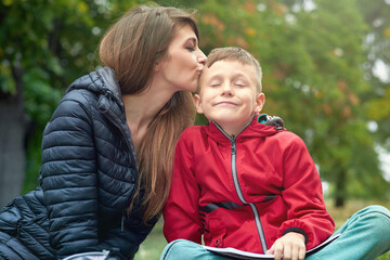 Young mother and a cute boy in a park, mum is kissing her son, rewarding his good work; he is writing in a book.Young female tutor mom teacher doing homework with her son outdoors. - obrazy, fototapety, plakaty