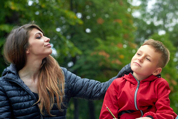 Young mother and a cute boy in a park, mum is kissing her son, rewarding his good work; he is...