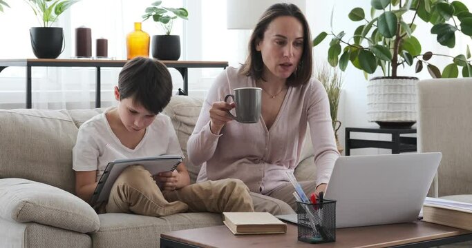 Mother helping son doing homework while drinking coffee and working on laptop at home