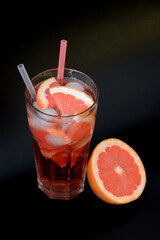 A tall glass of grapefruit juice with ice and pieces of ripe citrus on a black background.