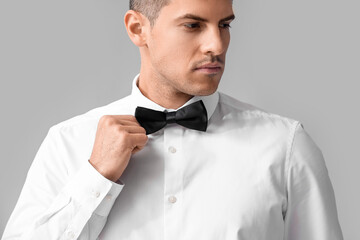 Fashionable young gentleman looking aside and adjusting bow tie on grey background