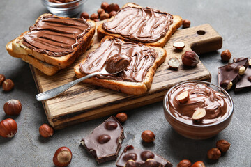 Wooden board of bread with chocolate paste and hazelnuts on grey background