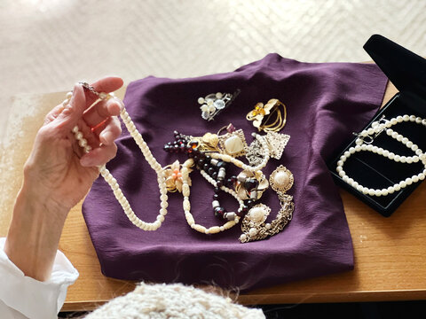 紫の布の上に置いたたくさんのジュエリーとパールのネックレスを持ち上げる高齢女性の手元