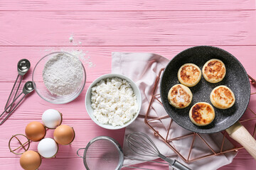 Frying pan with tasty cottage cheese pancakes and ingredients on pink wooden background