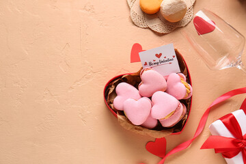 Box with tasty heart-shaped macaroons, greeting card, glass and gift on beige background
