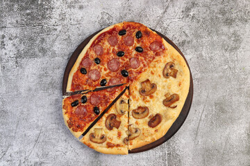Pizza in half with salami and olives, another - mushrooms with cheese sliced on a round wooden cutting board on a dark grey background. Top view, flat lay