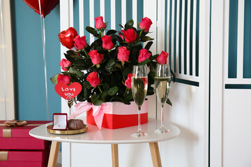 Box with roses, engagement ring and glasses of champagne on table near folding screen in room decorated for Valentine's Day
