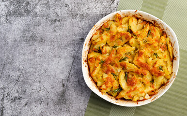 Potato casserole with cheese and rosemary in a white baking dish on a dark grey background. Top view, flat lay
