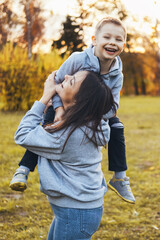 Mother holding her son on her shoulder having fun while playing a fun game in the park. Modern portrait of mother and boy enjoying free time in the park