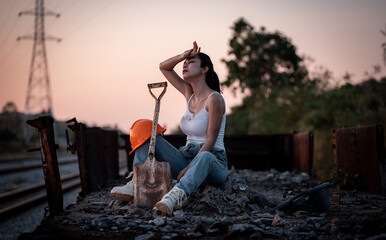 ฺBeautiful woman coal worker showed tried from work sitting on railway drinking water with orange...