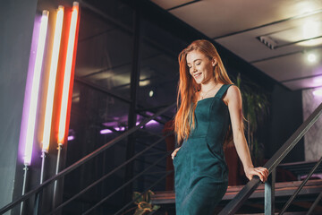 Caucasian redheaded woman in beautiful dress walking down from modern stairs in the restaurant. Elegant style. Beauty fashion model. Beautiful girl. Modern