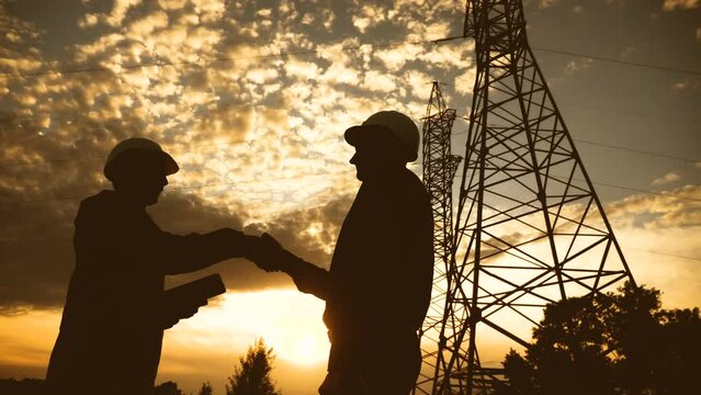 silhouette of an electricians engineers handshake next to an electrical support tower energy. electricity high teamwork tower support. electricals sunset engineers handshake teamwork electricity pylon